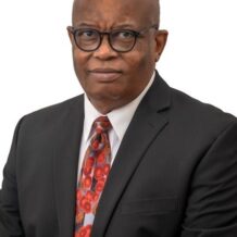 A man wearing glasses, a black suit, white shirt, and a colorful patterned tie, posing against a white background.