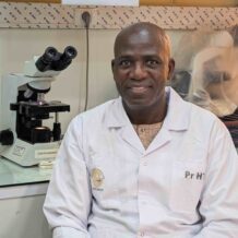 A man wearing a white lab coat sitting beside a microscope in a laboratory.