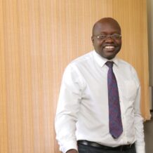 A smiling man in glasses, white shirt, and blue floral tie standing indoors, leaning on a chair.