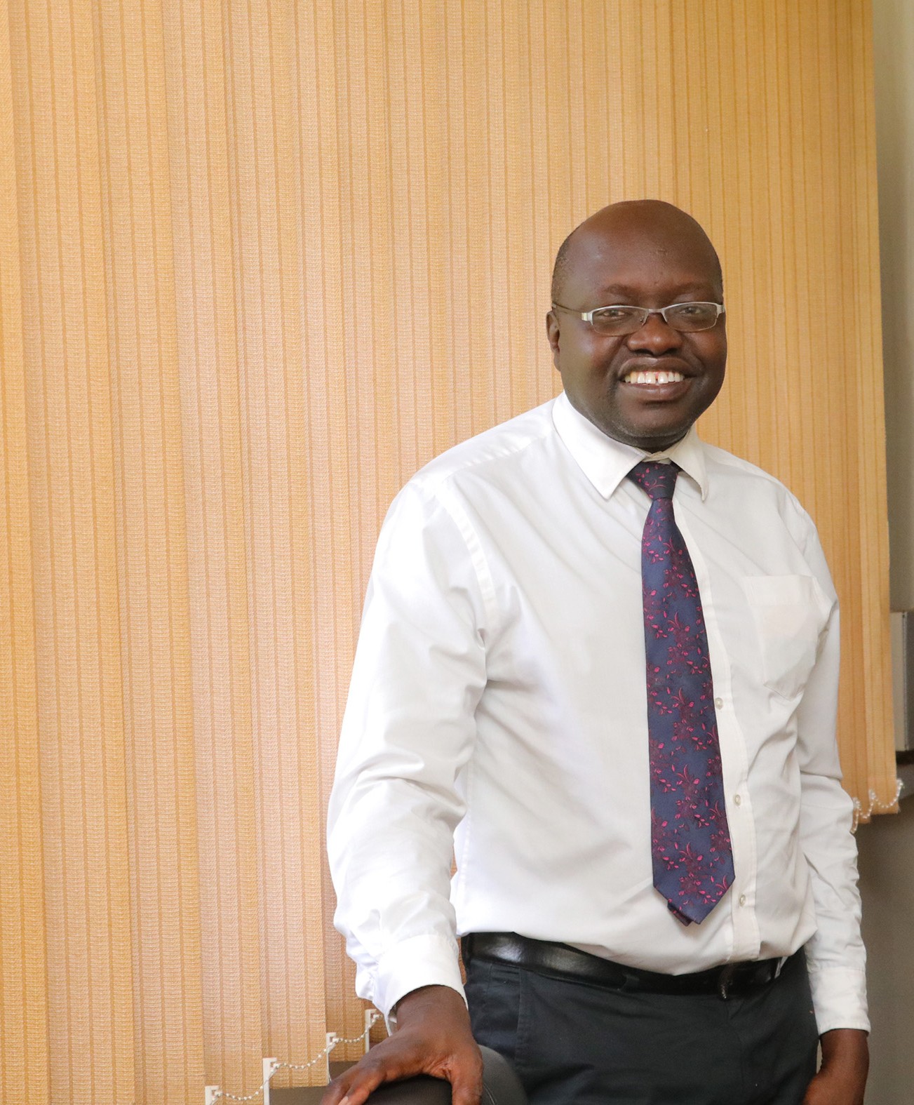 A smiling man in glasses, white shirt, and blue floral tie standing indoors, leaning on a chair.